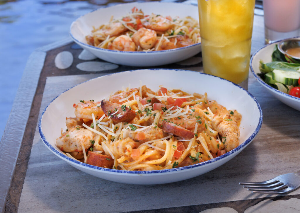 A close-up view of a creamy chicken, sausage, and shrimp pasta dish topped with parmesan cheese, on a table with other plates and a tropical drink.