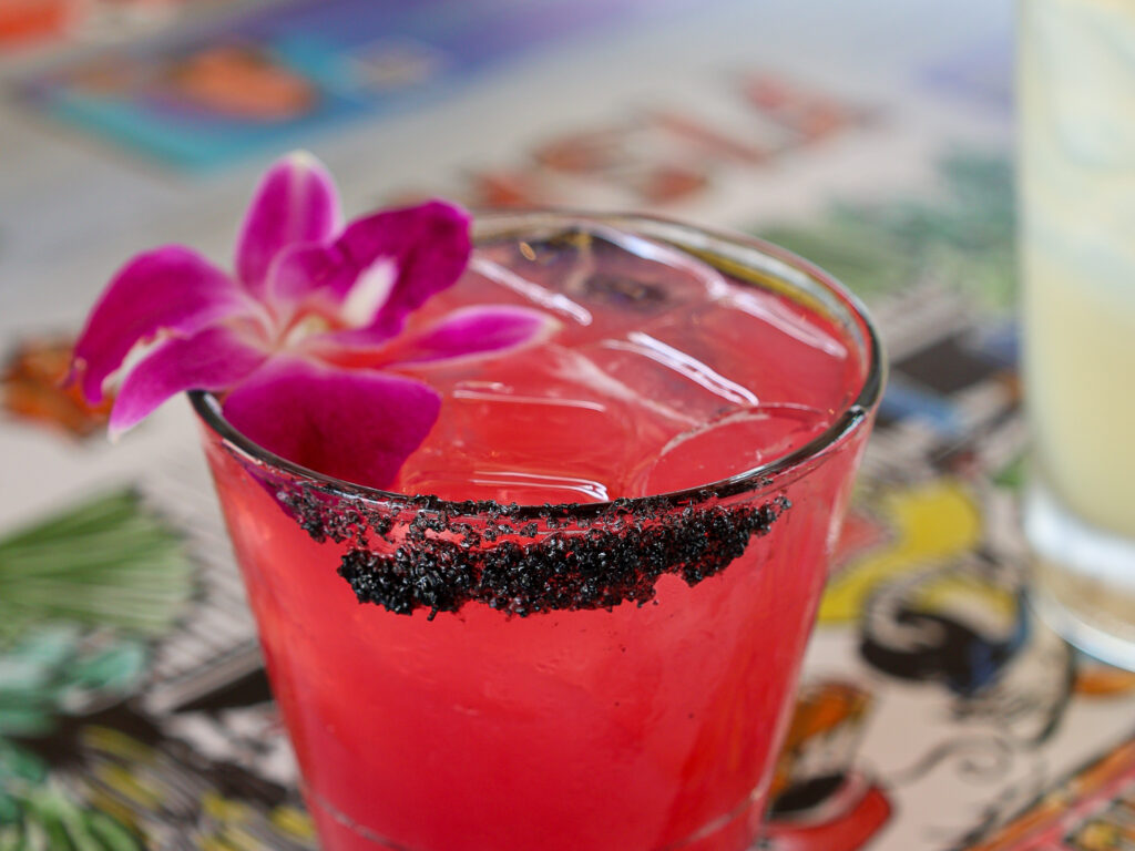 A vibrant pink cocktail served over ice in a lowball glass. The front rim is lined with coarse black volcanic salt, and the drink is garnished with a striking purple orchid flower. The glass rests on a brightly patterned tabletop with a blurred drink visible to the right.