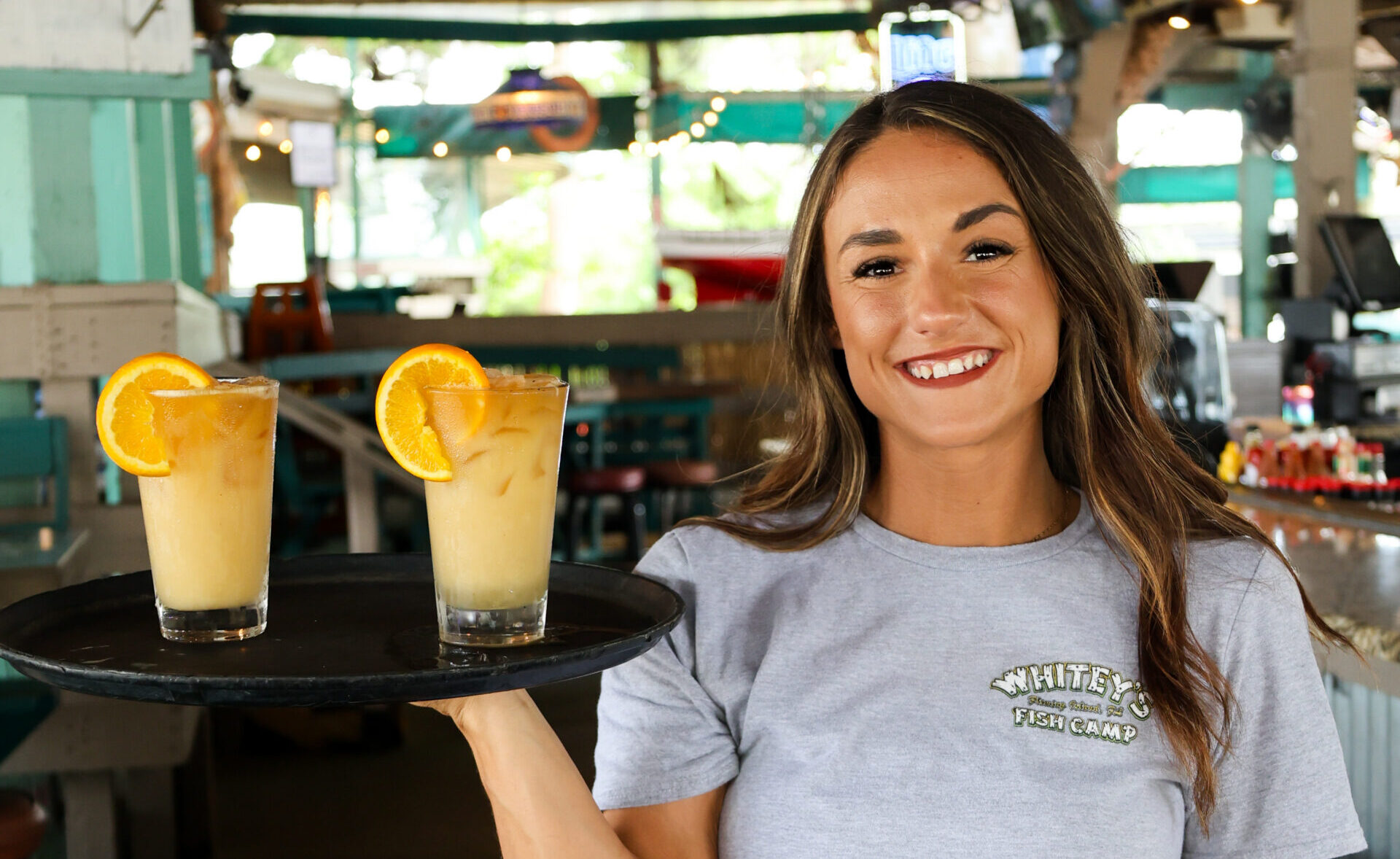 A smiling server at Whitey's Fish Camp holding a tray with two iced tropical cocktails garnished with orange slices.