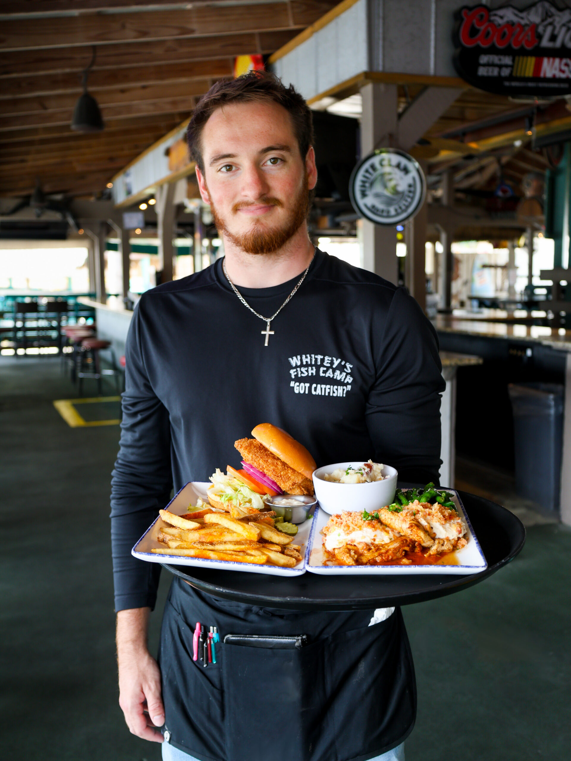 A server at Whitey's Fish Camp holding a tray filled with a fried fish sandwich, french fries, and a seafood entrée in an open-air restaurant setting.