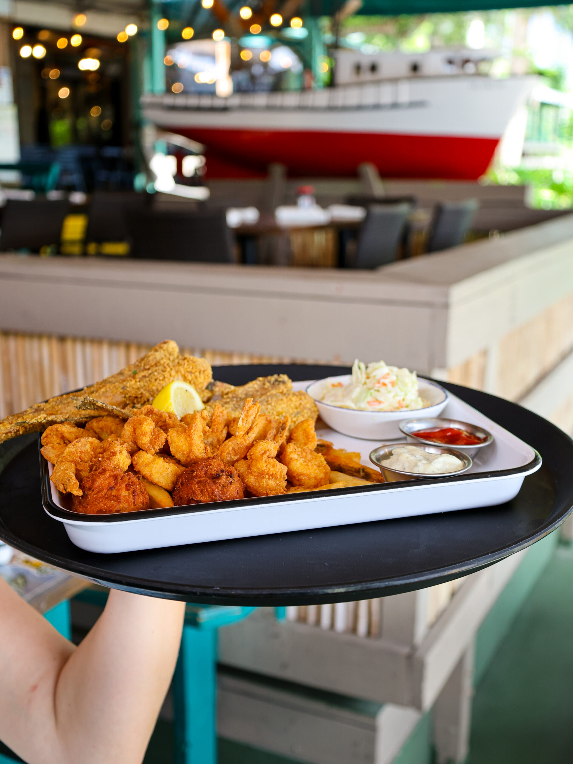 A fried seafood platter with fried shrimp, fish, hushpuppies, coleslaw, and dipping sauces on a server's tray at a waterfront restaurant.