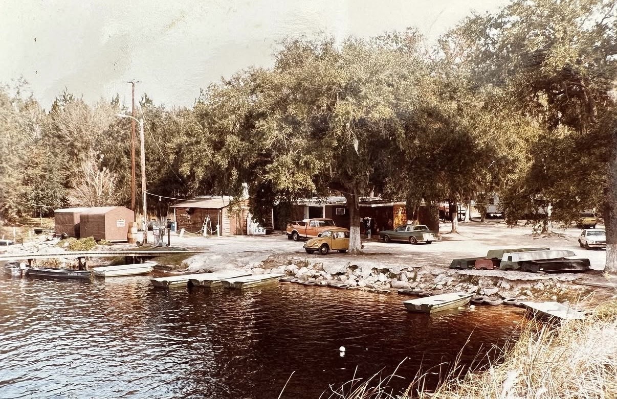 A vintage photograph of an old fish camp on a riverbank with rustic wooden buildings, classic cars like a VW Beetle, and small fishing boats moored along the shore.