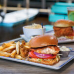 A close-up of a seasoned fish sandwich on a brioche bun, layered with red onion, tomato, and lettuce, served on a white rectangular plate alongside a pile of french fries and a small cup of tartar sauce. In the blurred background, another sandwich, a bright red cocktail, and a waterfront dock setting are visible.