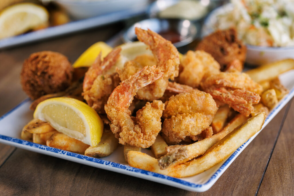 A plate of fried shrimp, french fries, and hush puppies garnished with lemon wedges, with coleslaw and dipping sauces in the background.