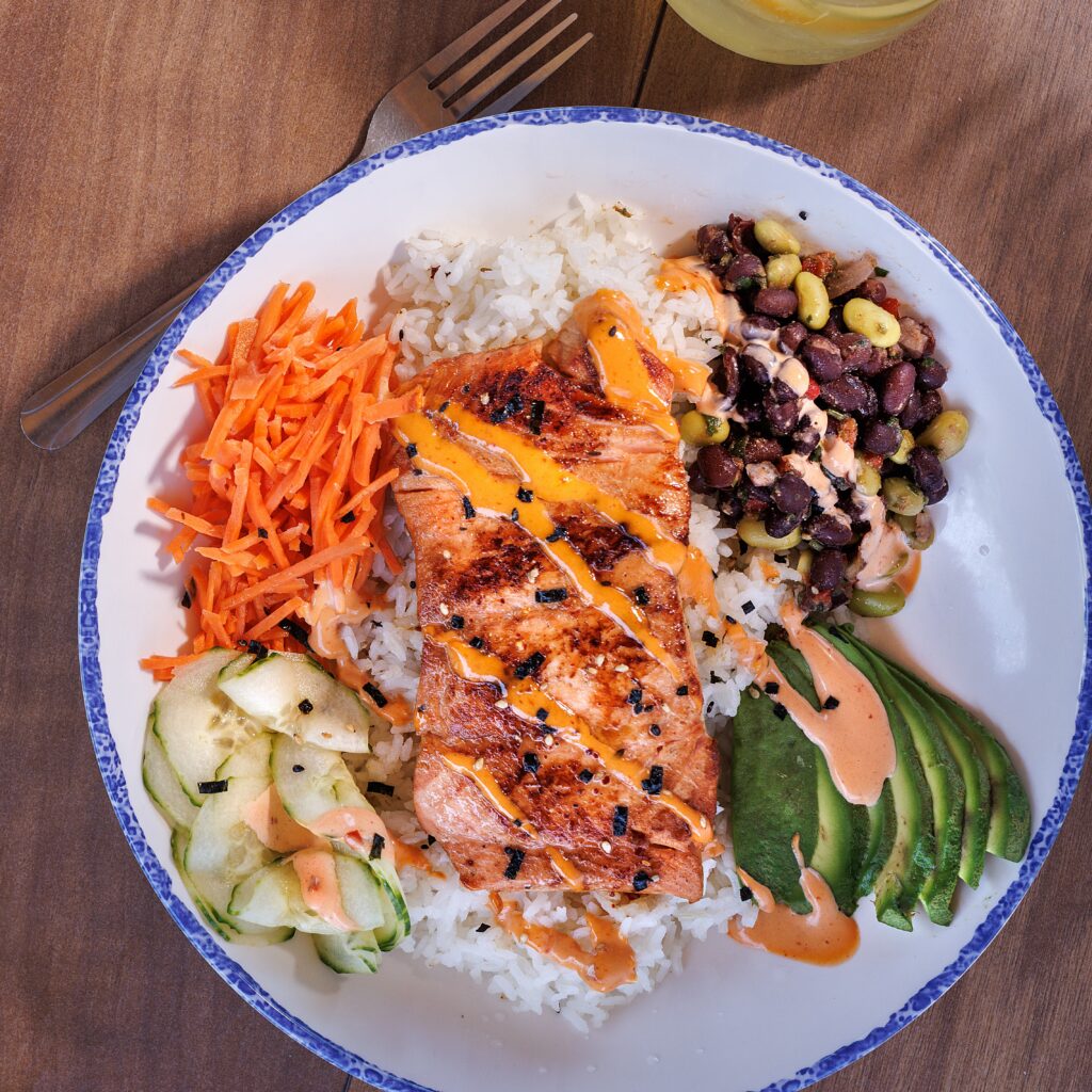 A grilled salmon poke bowl with white rice, avocado, shredded carrots, cucumbers, and a black bean salad on a wooden table.
