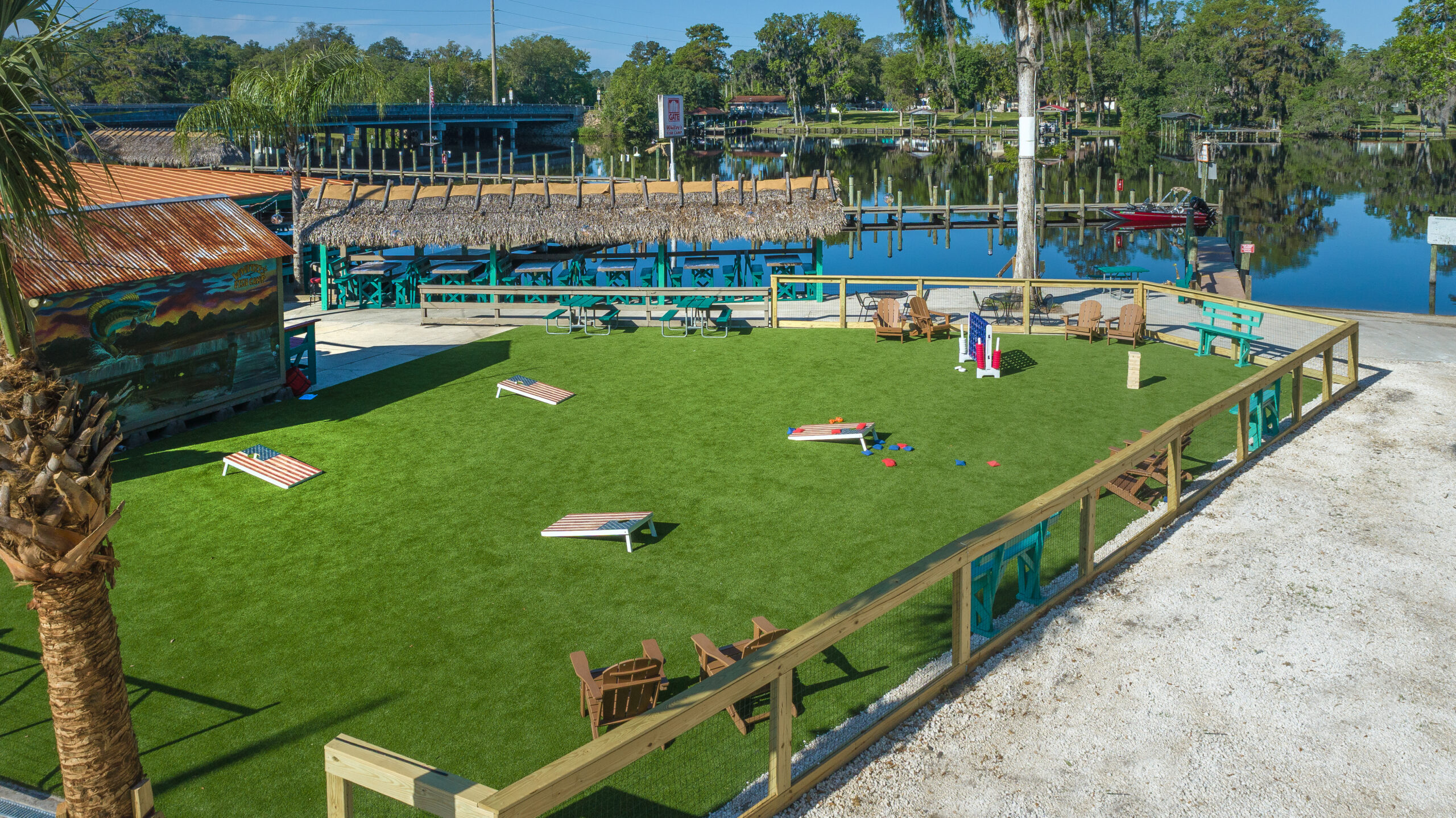 A high-angle shot of an outdoor recreational area at a waterfront venue. A fenced-in artificial turf lawn features several lawn games, including corn hole boards painted like American flags and a giant Connect Four set. In the background, a covered patio with a thatched roof, picnic tables, a wooden boat dock, and a calm river under a clear blue sky are visible.