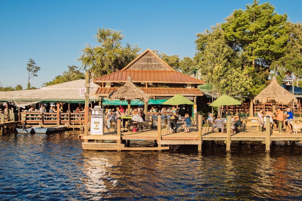 A sunny, wide shot of a bustling waterfront restaurant and tiki bar viewed from the water. Patrons are dining on a wide wooden deck lined with green umbrellas and thatched palapa roofs. The main open-air building features a copper-colored metal roof, and lush green trees surround the property under a clear blue sky.