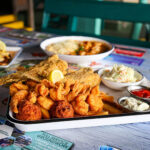 A close-up view of a large fried seafood platter with shrimp, fish, hushpuppies, fries, coleslaw, and dipping sauces on a restaurant table.