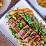 An overhead view of a seared ahi tuna salad platter with arugula, spicy mayo, green onions, and black sesame seeds.