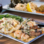 Grilled steak topped with shrimp and a creamy sauce, served with red potato mashed potatoes and green beans on a rectangular plate, with a glass of red wine and additional entrées in the background.