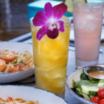 A close-up view of a tall yellow cocktail garnished with a purple orchid, surrounded by plates of pasta and a salad on a restaurant table.