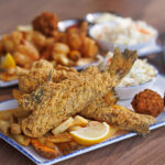 A close-up view of a whole fried fish platter with french fries, hushpuppies, a lemon wedge, and coleslaw on a restaurant table.