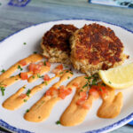 A close-up view of two pan-seared crab cakes on a plate with lines of creamy orange sauce, diced tomatoes, and a lemon wedge.