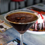 A close-up view of an espresso martini garnished with three coffee beans, with a blurred chocolate dessert in the background.