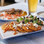 A plate of fried green tomatoes topped with crumbled cheese and sauce, served next to a pint of beer and crab cakes at a waterfront restaurant table.