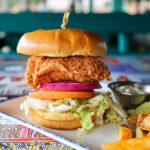 A tall fried fish sandwich with lettuce, tomato, and red onion on a bun, served with french fries and tartar sauce at a restaurant table.