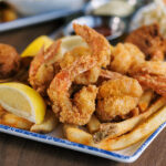 A plate of fried shrimp, french fries, and hush puppies garnished with lemon wedges, with coleslaw and dipping sauces in the background.