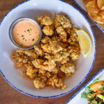 An overhead view of a bowl of crispy fried gator tail bites with an orange dipping sauce and a lemon wedge.