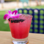 A close-up view of an iced pink cocktail with a black salt rim and a purple orchid garnish, sitting on an outdoor wooden table.