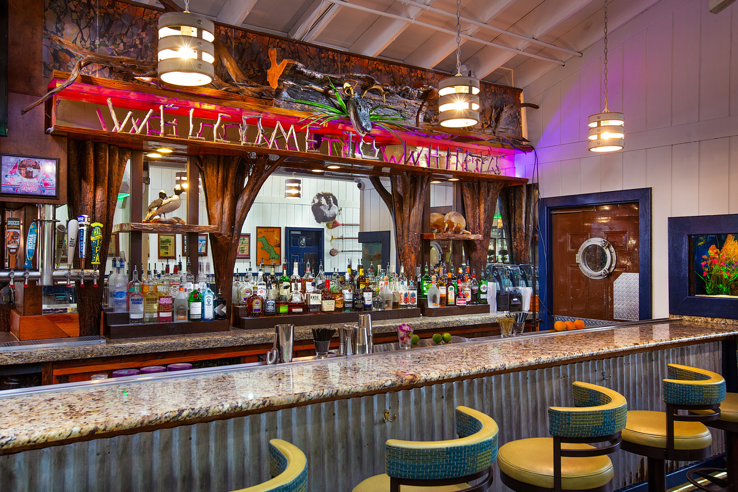 A bar with a granite counter, bar stools, and a mirrored back wall displaying bottles, taps, and decorative elements under warm lighting.