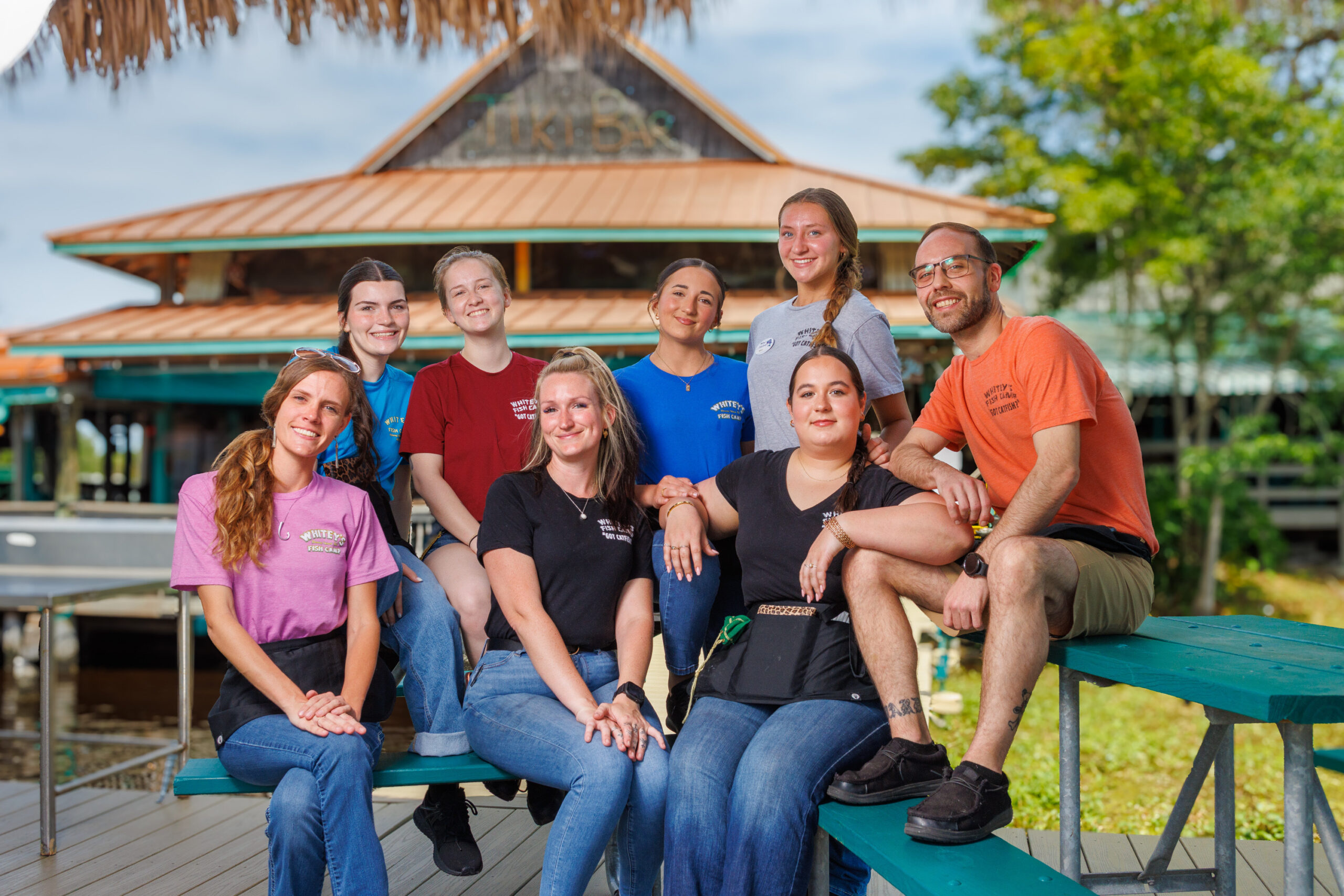 A group of eight smiling restaurant employees posing together outdoors on a turquoise picnic table. They are wearing casual t-shirts in various colors, most with a "Whitey's Fish Camp" logo, and several wear black server aprons. In the background, a large waterfront "Tiki Bar" building with a metal roof is visible under a slightly overcast sky.