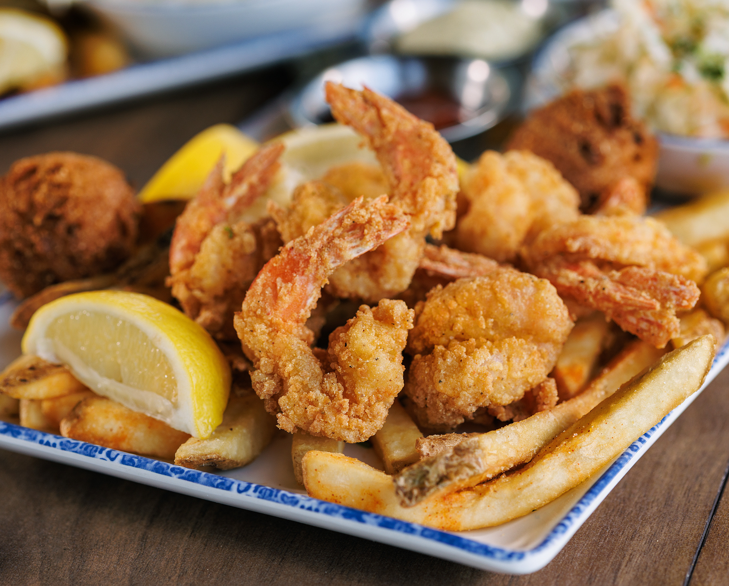 A plate of fried shrimp, french fries, and hush puppies garnished with lemon wedges, with coleslaw and dipping sauces in the background.