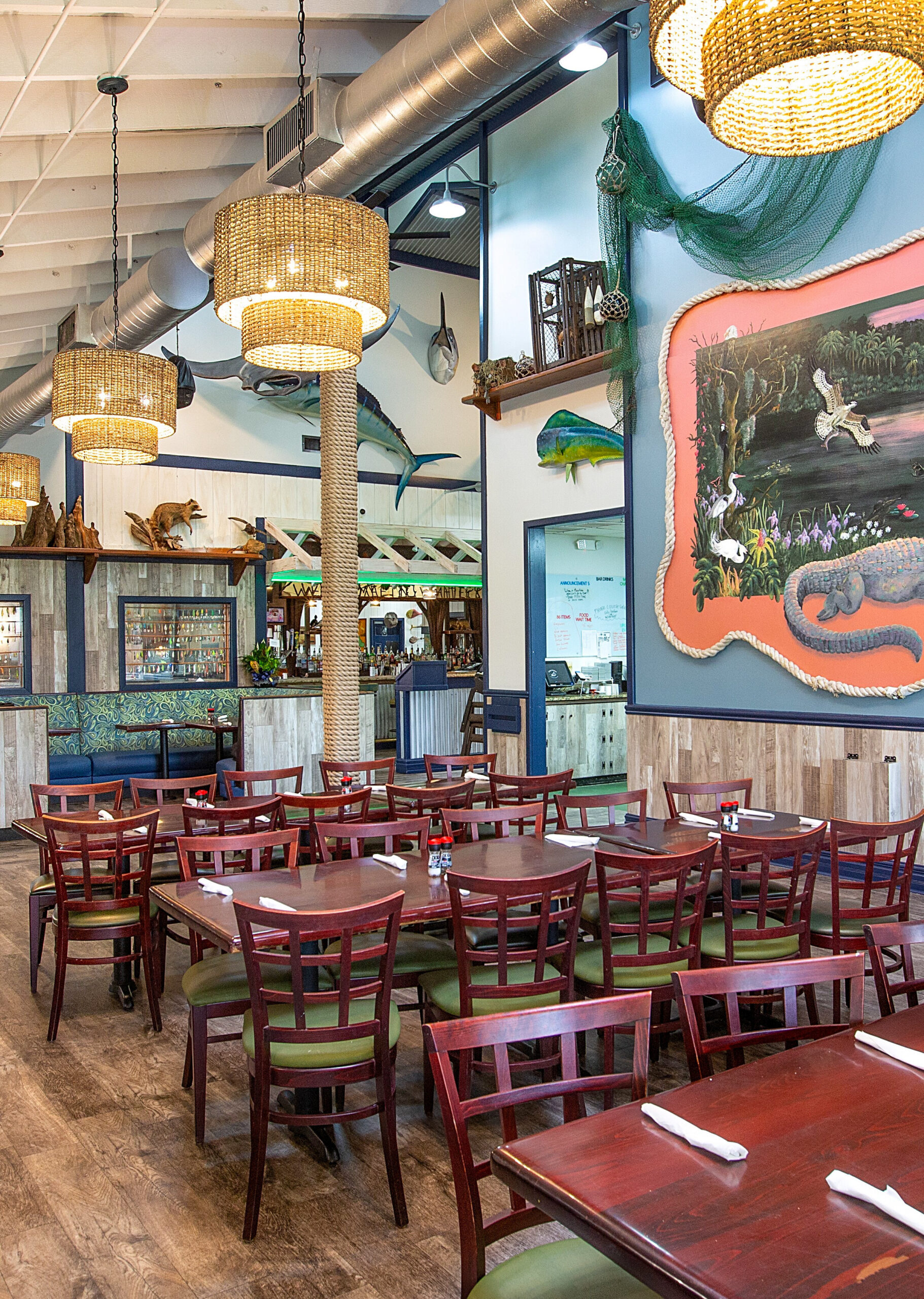 Interior of a coastal-themed restaurant dining room with wood tables, woven basket chandeliers, and Florida-inspired wildlife decor on the walls.
