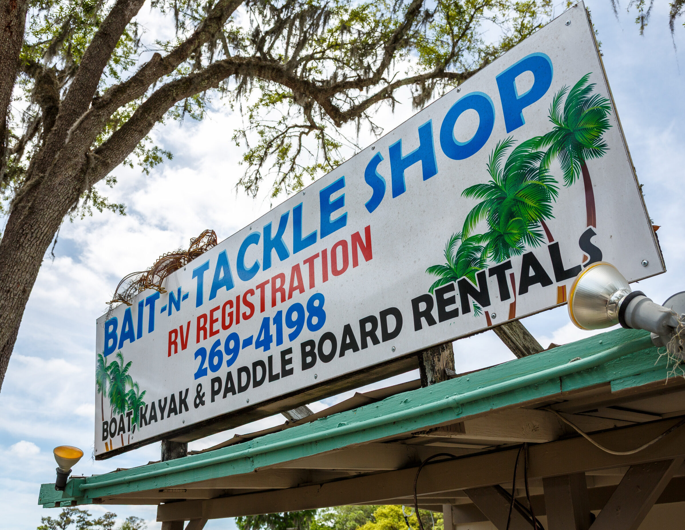 A sign for a bait and tackle shop with palm tree graphics, offering RV registration, and boat, kayak, and paddle board rentals. Trees and sky in the background.