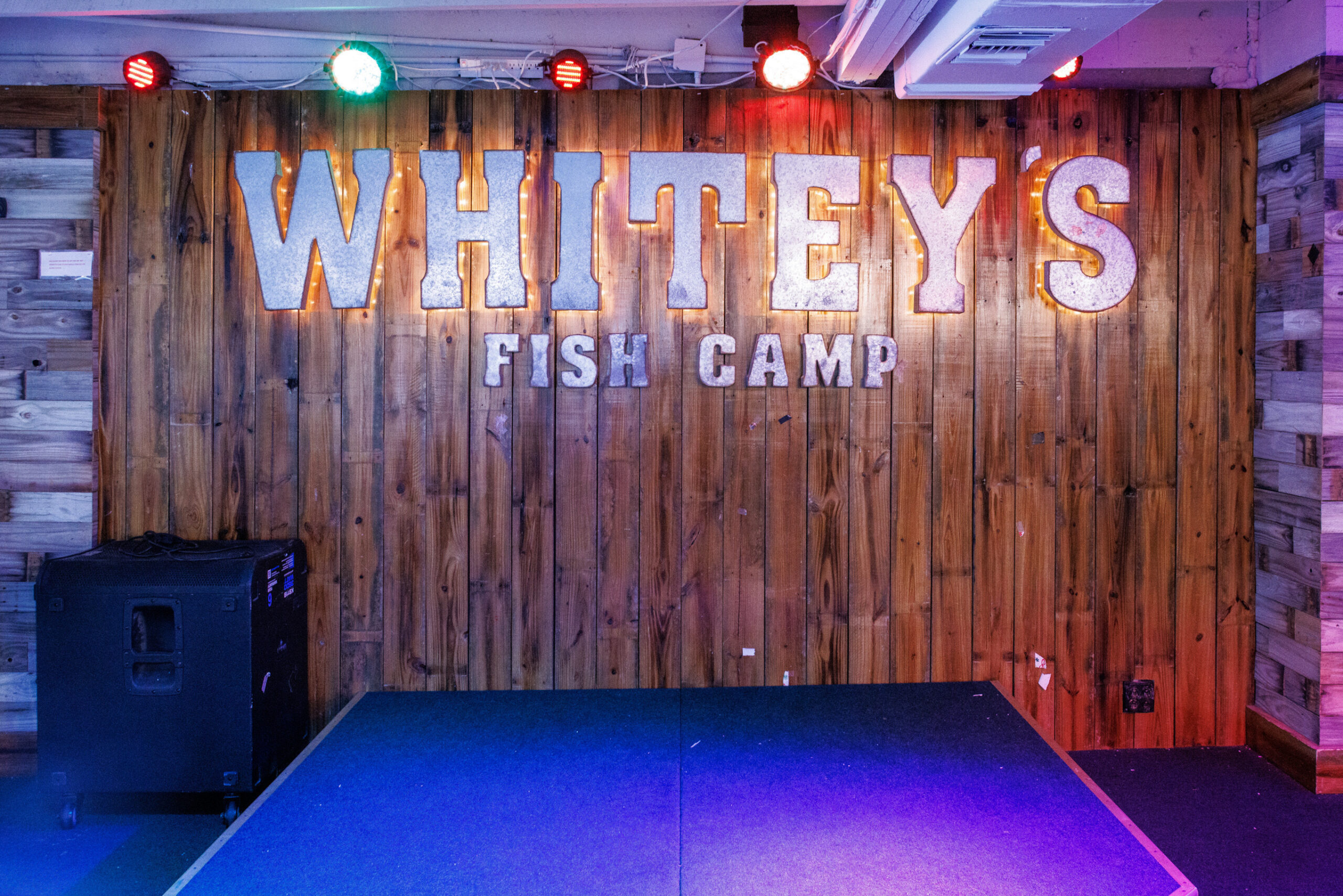 Wood-paneled stage with vibrant lighting displays "Whitey's Fish Camp" in bold letters. Warm, rustic ambiance with red, green, and white lights overhead.