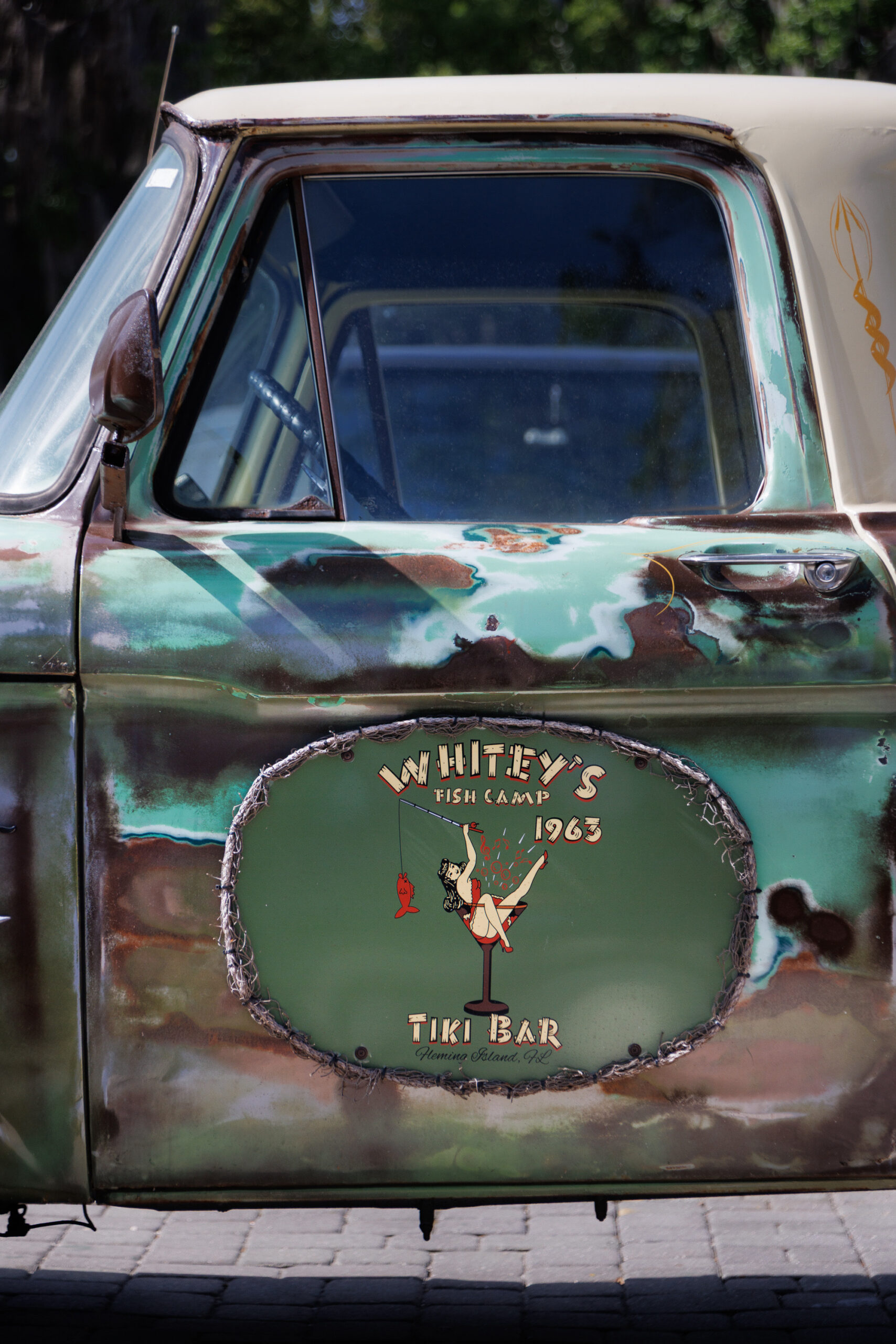 Close-up of a weathered vintage pickup truck door with a green and rusted finish, featuring a "Whitey's Fish Camp 1963 Tiki Bar" logo painted on the side.