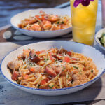 A close-up view of a chicken, sausage, and shrimp pasta dish topped with parmesan cheese and diced tomatoes, served on a restaurant table with a tropical drink in the background.