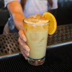 A close-up view of a hand serving a tall, iced tropical cocktail with a frothy top, spiced dust, and an orange slice garnish at a bar.