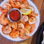 A top-down view of seasoned peel-and-eat shrimp arranged with cocktail sauce and a lemon wedge in a blue-rimmed white bowl.