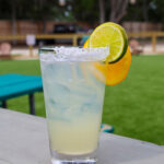 A tall iced margarita in a pint glass with a salted rim, garnished with an orange and lime slice, sitting on an outdoor table next to a green lawn.