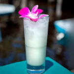 A tall, icy white cocktail garnished with a bright pink orchid flower, sitting on a teal surface with a blurry water background.