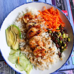 A top-down view of a grilled shrimp bowl with white rice, avocado, cucumbers, shredded carrots, and an edamame bean salad.