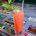 A tall pink cocktail with ice, a straw, and a fresh mint garnish sitting on a decorated restaurant table.