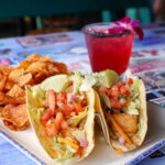 two tacos with fried fish, lettuce, tomatoe and pico. Drink in background.